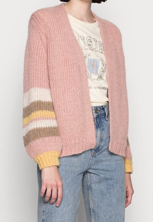 Femme portant un cardigan en maille épaisse rose avec des manches rayées, un t-shirt graphique et un jean bleu taille haute sur un fond uni.