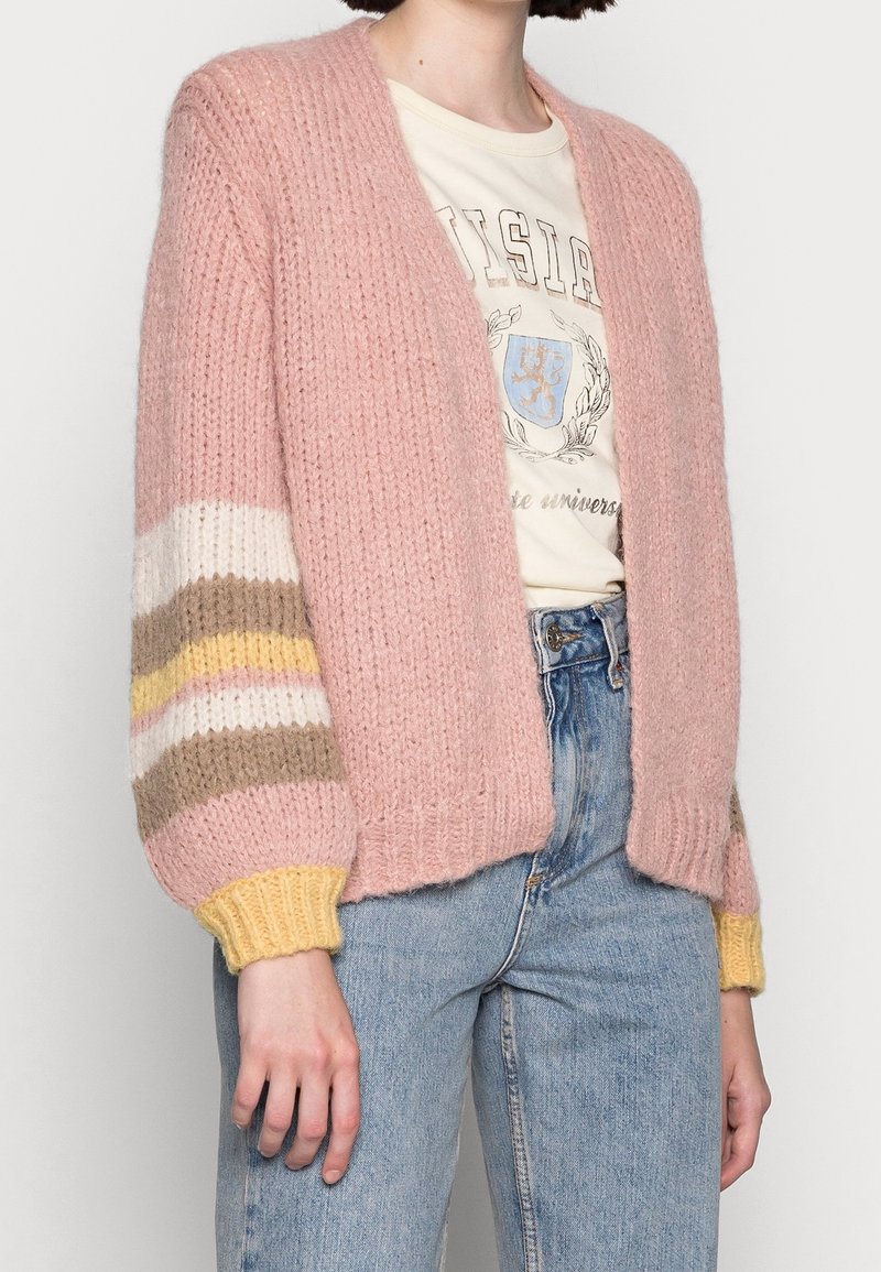 Femme portant un cardigan en maille épaisse rose avec des manches rayées, un t-shirt graphique et un jean bleu taille haute sur un fond uni.