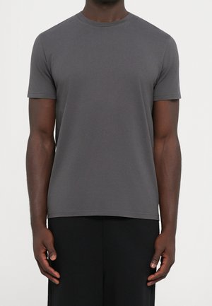 Hombre que lleva una camiseta gris oscuro sencilla de manga corta y cuello redondo y pantalones negros, de pie frente a un fondo blanco.