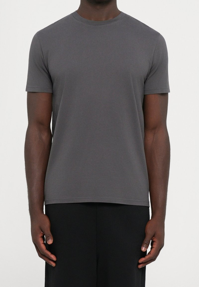 Homme portant un t-shirt gris foncé uni à manches courtes et col rond, et un pantalon noir, debout devant un fond blanc.
