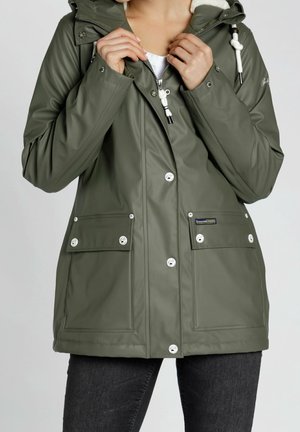 Personne portant une veste de pluie à capuche vert olive avec des cordons blancs et des poches avant, en train de fermer la fermeture éclair près du col.