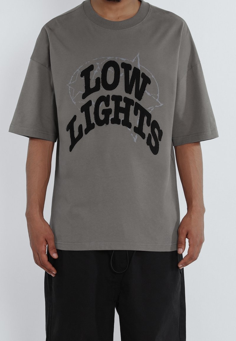 T-shirt oversize gris avec un texte noir audacieux "LOW LIGHTS" et un contour graphique subtil. Manches courtes, col rond, tissu doux.
