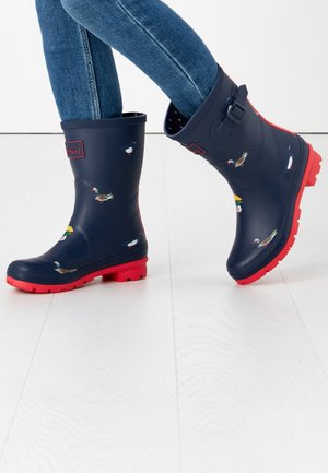 Granatowe gumowe kalosze z czerwonym podeszwa, zdobione ilustracjami kaczek i szczegółem w postaci klamry z boku. Gładka faktura, wysokość do połowy łydki.