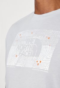 Ljusgrå bomullströja med en vit topografisk kartdesign med orange detaljer och märketext: "THE NORTH FACE" och "NEVER STOP EXPLORING."