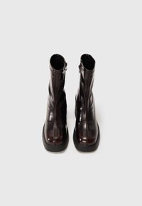Par de botas de cuero marrón oscuro pulido hasta el tobillo, con puntera cuadrada y suelas gruesas de goma negra, sobre un fondo neutro.