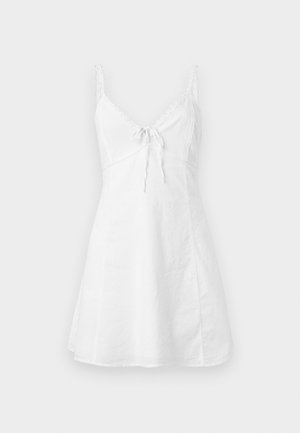 Vestido de verano blanco sin mangas con finas tiras de encaje y detalle de lazo atado en la parte delantera sobre un fondo liso.