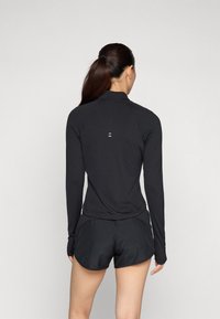 Crni dugos manjim rukavima sportski top s polu-visokim ovratnikom, s diskretnim logotipom na leđima, u kombinaciji s crnim sportskim kratkim hlačama.