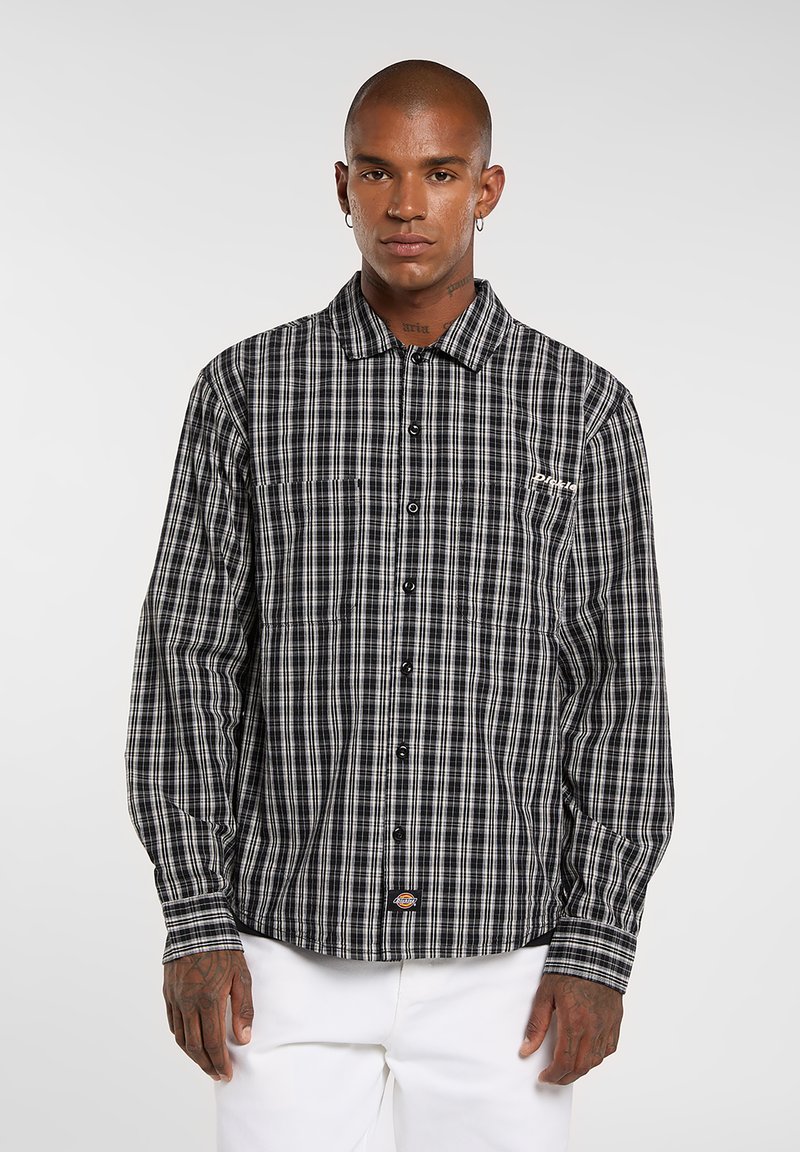 Camicia a quadri bianca e nera a maniche lunghe con colletto, bottoni sul davanti e taschino sul petto. Presenta un tessuto testurizzato e dettagli di cuciture chiare.