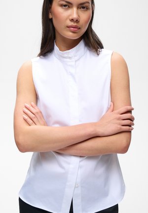 Femme aux longs cheveux portant une chemise blanche sans manches à boutons, debout les bras croisés devant un fond clair et uni.