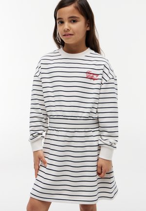 Ung pige iført en hvid og sort stribet langærmet kjole med rødt broderet "Tommy Hilfiger" logo på brystet, poserer med hænderne ned.