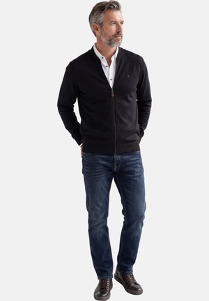 Mann mit grauem Haar und Bart, der einen schwarzen Pullover mit Reißverschluss, ein weißes Hemd, blaue Jeans und dunkelbraune Schuhe trägt, steht mit den Händen hinter dem Rücken.