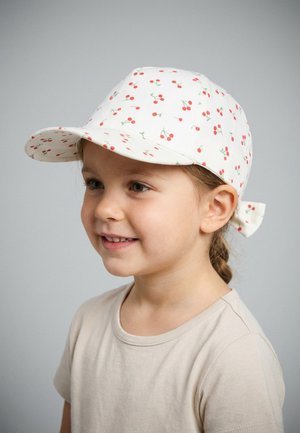 Young girl smiling, wearing a white cap with small red cherry patterns and a light beige shirt, facing right against a plain grey background.