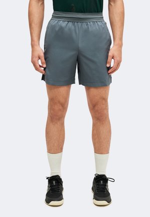 Mann trägt graue sportliche Shorts, weiße Kniestrümpfe und schwarze Turnschuhe, steht vor einem hellen Hintergrund.