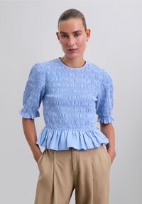 Femme avec les cheveux plaqués en arrière, portant un blouse à manches bouffantes texturée bleu clair et un pantalon plissé taille haute beige, les mains dans les poches.