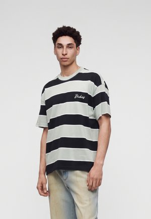 Jovem usando uma t-shirt larga às riscas horizontais preto e branco e jeans azul claro, parado contra um fundo claro simples.