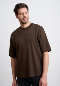 Brązowa koszulka z krótkim rękawem wykonana z miękkiej bawełny, z klasycznym okrągłym dekoltem i luźnym krojem, w połączeniu z ciemnymi dżinsami.