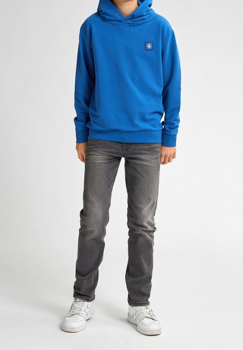 Hoodie en coton bleu avec une poche frontale et un patch de logo, associé à un jean gris et des baskets blanches. Texture lisse et coupe décontractée.