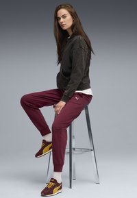 Dunkelgrauer Hoodie mit lässiger Passform, burgunderfarbene Jogginghose mit gerippten Bündchen und kastanienbraune Sneaker mit gelben Akzenten. Auf einem Hocker sitzend.