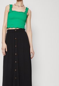 Vihreä, smokkikuvioinen lyhyt toppi röyhelöhihnoilla, yhdistetty mustan, napillisen maksi-hameen kanssa, jossa on teksturoitu kangas.