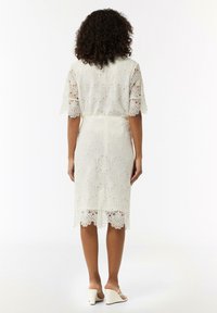 Robe en dentelle blanc cassé avec un motif floral, manches courtes, longueur genoux et bords festonnés, associée à des sandales à talons blanches.