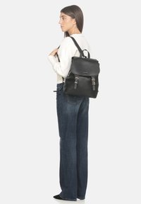 Zaino in pelle nera con patta arrotondata, accessori in argento e spallacci regolabili. Indossato sulla schiena di una persona in abbigliamento casual.