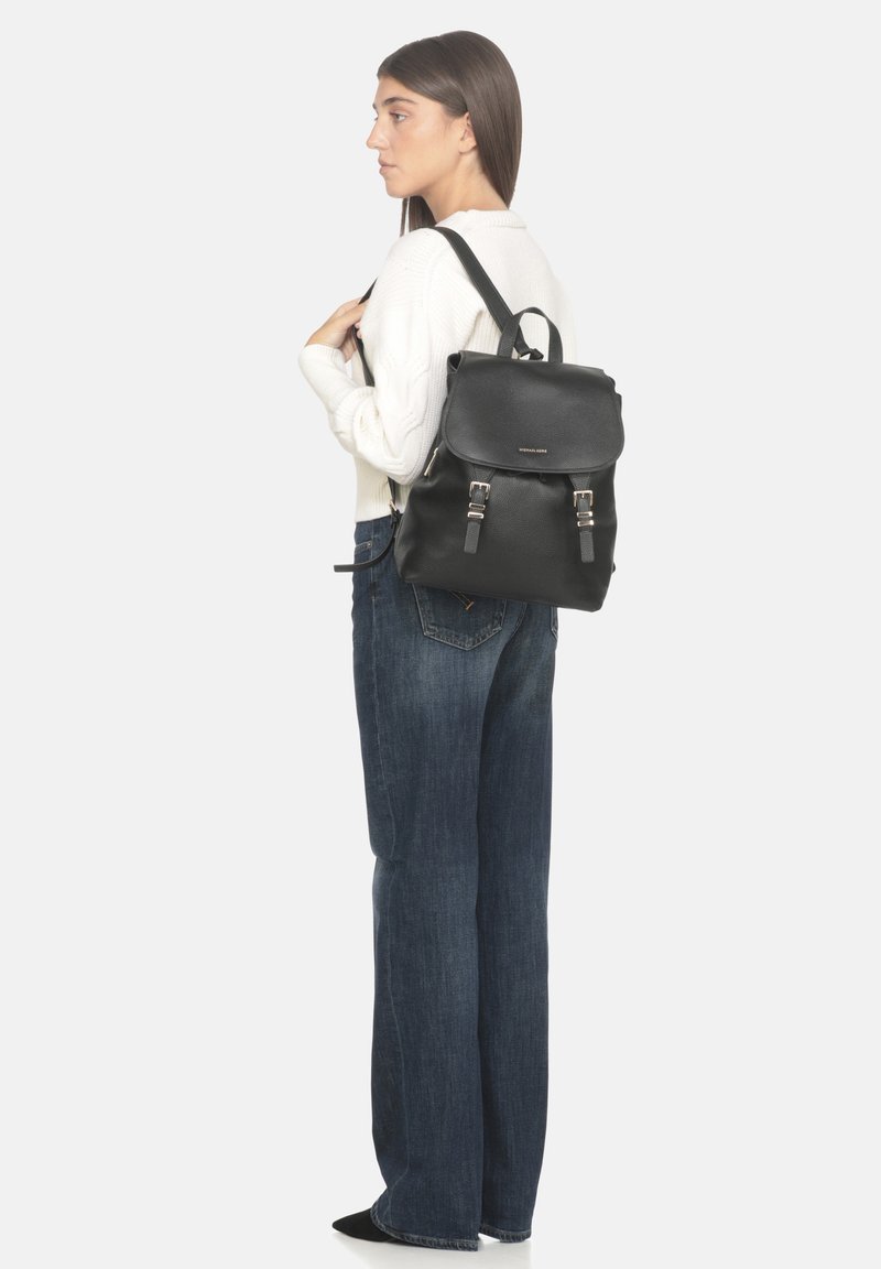 Zaino in pelle nera con patta arrotondata, accessori in argento e spallacci regolabili. Indossato sulla schiena di una persona in abbigliamento casual.