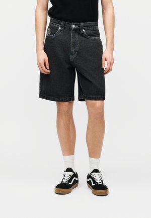 Άνδρας που φοράει μαύρα τζιν σορτς, μαύρο μπλουζάκι, λευκές κάλτσες και μαύρα αθλητικά παπούτσια με λευκά κορδόνια, στέκεται μπροστά σε απλό φόντο.