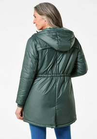 Die grüne isolierte Jacke hat eine Kapuze, einen verstellbaren Bund und eine gesteppten Textur. Der Stoff hat eine glatte, glänzende Oberfläche. Darunter werden blaue Leggings getragen.