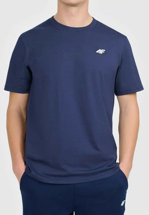 Mężczyzna w granatowym krótkim T-shircie i pasujących spodniach z małymi białymi logo „4F” na klatce piersiowej i udzie, z rękami w kieszeniach.