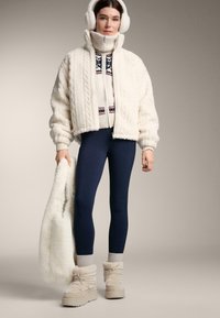 Beżowa kurtka z futerkiem z ekoskóry, kremowy sweter w warkocze z wzorzystymi akcentami, granatowe legginsy, kremowe nauszniki oraz beżowe zimowe buty.