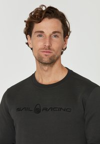 Ciemnoszara bluza z miękkiego materiału, z okrągłym dekoltem, długimi rękawami oraz haftowanym logo "SAIL RACING" na piersi.