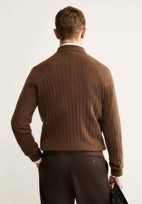 Maglione marrone a coste con colletto alto, indossato sopra pantaloni marrone scuro. L'outfit presenta un tessuto testurizzato e dettagli bianchi delicati al colletto.