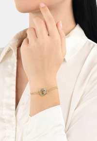 Guldkedjearmband med silverfärgade sammanlänkade cirkulära accenter, bärs på en hand mot en vit blus, vilket framhäver den delikata designen.