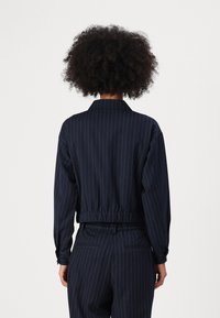 Donkerblauwe gestreepte jas met een elastische tailleband, lange mouwen en een klassieke kraag. Voorzien van verticale witte streepjes voor textuur.