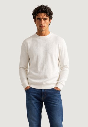 Mand med krøllet mørkt hår iført en hvid tekstureret sweater og blå jeans, står med hænderne i lommerne mod en ensfarvet baggrund.