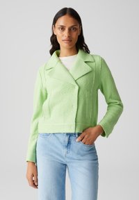 Giacca corta verde chiaro in tessuto texture con colletto a revers, chiusura a bottoni e due tasche frontali, abbinata a jeans azzurri.