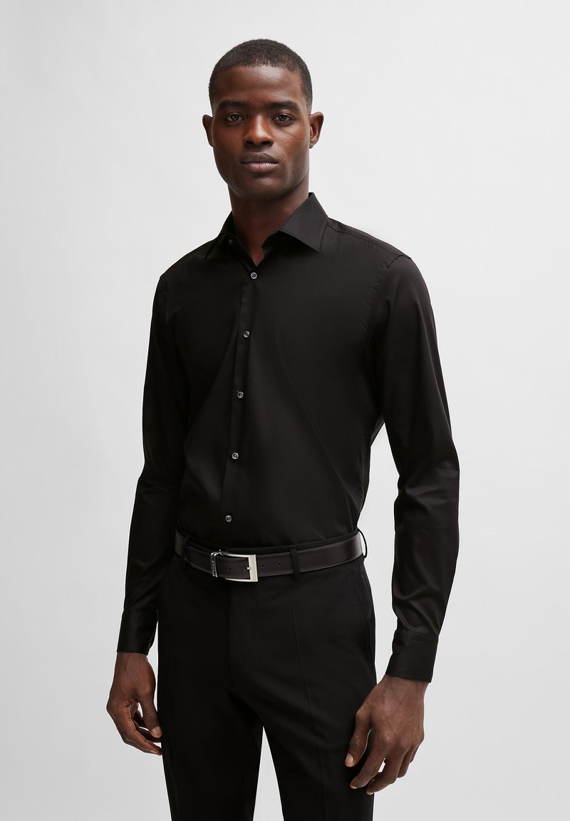 Camisa negra de manga larga con cuello clásico, cierre frontal de botones y tela suave; combinada con pantalones negros y un cinturón marrón con herrajes plateados.