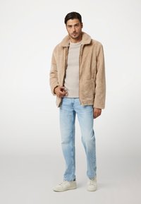 Giacca in pelliccia sintetica beige con colletto, tasche frontali e vestibilità rilassata, abbinata a jeans azzurri e sneakers bianche.