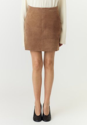 Kvinde iført en tanfarvet ruskind mininederdel, cremefarvet langærmet sweater og sorte lukkede hæle, stående mod en ensfarvet baggrund.