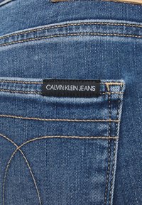 Acercamiento al bolsillo trasero de unos jeans de mezclilla azules con costuras visibles y una etiqueta negra que dice "Calvin Klein Jeans."