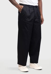 Zwarte geplooide broek met zijzakken en taps toelopende pijp, gemaakt van gladde stof, gecombineerd met beige sneakers.