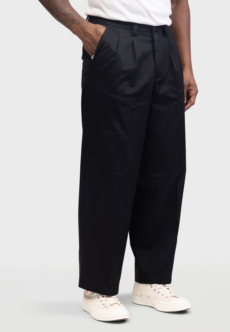 Zwarte geplooide broek met zijzakken en taps toelopende pijp, gemaakt van gladde stof, gecombineerd met beige sneakers.