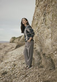 Szary sweter w kratę na granatowym tle, w zestawieniu z jasnoszarymi spodniami i brązowymi sneakerami. Model stoi na kamienistej plaży.