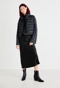 Chaleco acolchado negro sobre un suéter gris oscuro, combinado con una falda midi de satén negra y botas de tobillo negras con tacón. Texturas suaves, diseño en capas.