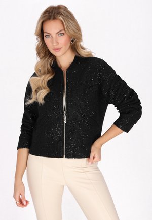 Giacca nera con zip realizzata in un tessuto testurizzato e luccicante. Presenta un collo rotondo e maniche a tre quarti. Abbinata a pantaloni beige aderenti.