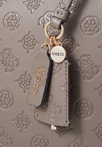 Grå syntetisk håndtaske med præget mønster, guld hardware detaljer og et aftageligt mærke med mærkenavnet "GUESS."
