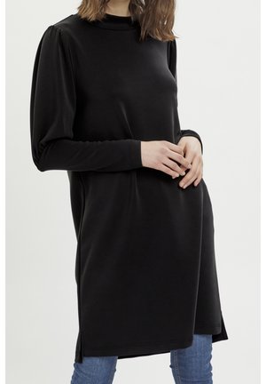 Femme portant une longue robe noire avec des manches longues bouffantes, debout les mains jointes sur un jean bleu sur un fond blanc.