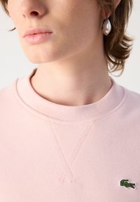 Ljusrosa sweatshirt med ribbad krage, texturerat tyg och grön krokodil-logotyp på bröstet. Närbild på halsringning och örhänge.