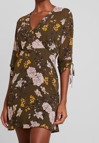 Florales Wickelkleid in Olivgrün mit pinken und gelben Blumen. Weicher Stoff, V-Ausschnitt, dreiviertellange Ärmel mit Bindebändern.