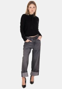 Giovane donna in piedi con le mani nelle tasche, che indossa un top a maniche lunghe nero, jeans larghi grigi con risvolti e tacchi neri.